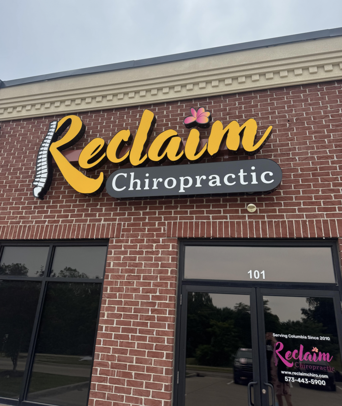 Exterior building signage for Reclaim Chiropractic. Large dimensional letters in gold script spell "Reclaim" with a spine graphic integrated into the letter R and a pink flower accent above the letter i. Below, a dark rounded rectangle panel displays "Chiropractic" in white serif lettering. The building is red brick, suite 101.