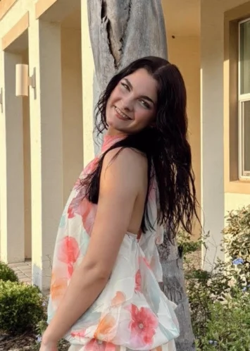 A young woman with dark hair smiles at the camera. Her head is tilted and she's looking over her left shoulder while outside. Taylor is a team member at Reclaim Chiropractic in Columbia, MO.